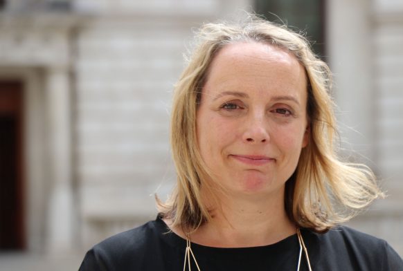 A women seen from the shoulders up smiling and wearing black standing in front of a stone building