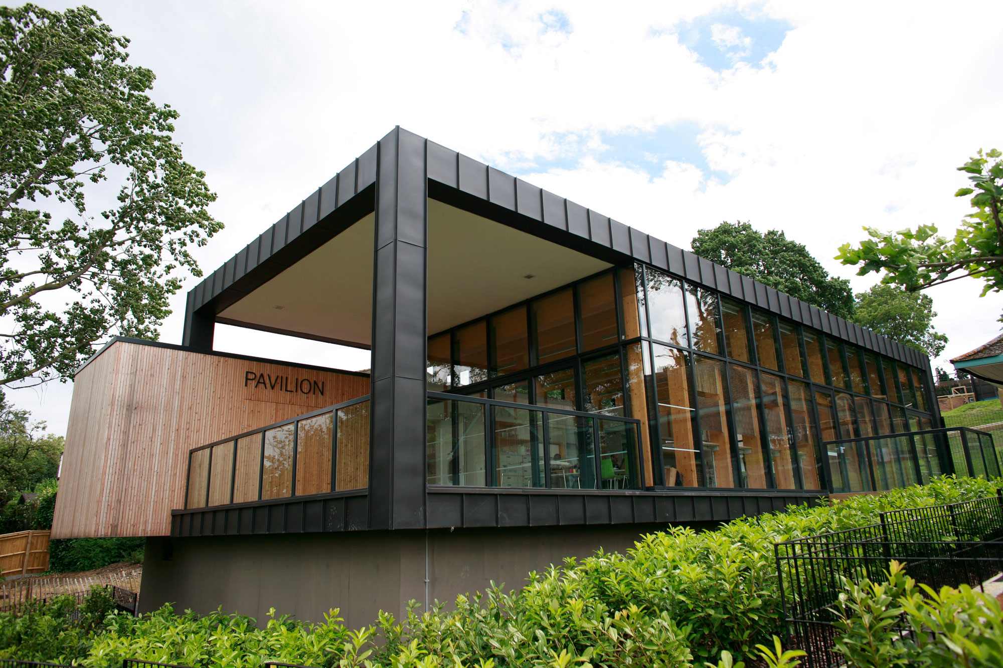 A square building as seen from outside and slightly below at an angle. The end closest to the camera is open to a balcony, with a glass walled internal room behind it. There are trees and shrubs surrounding it