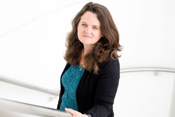 A woman is seen from the waist up standing on a staircase below the camera, she is holding on to the stair railing and the backdrop is bright white