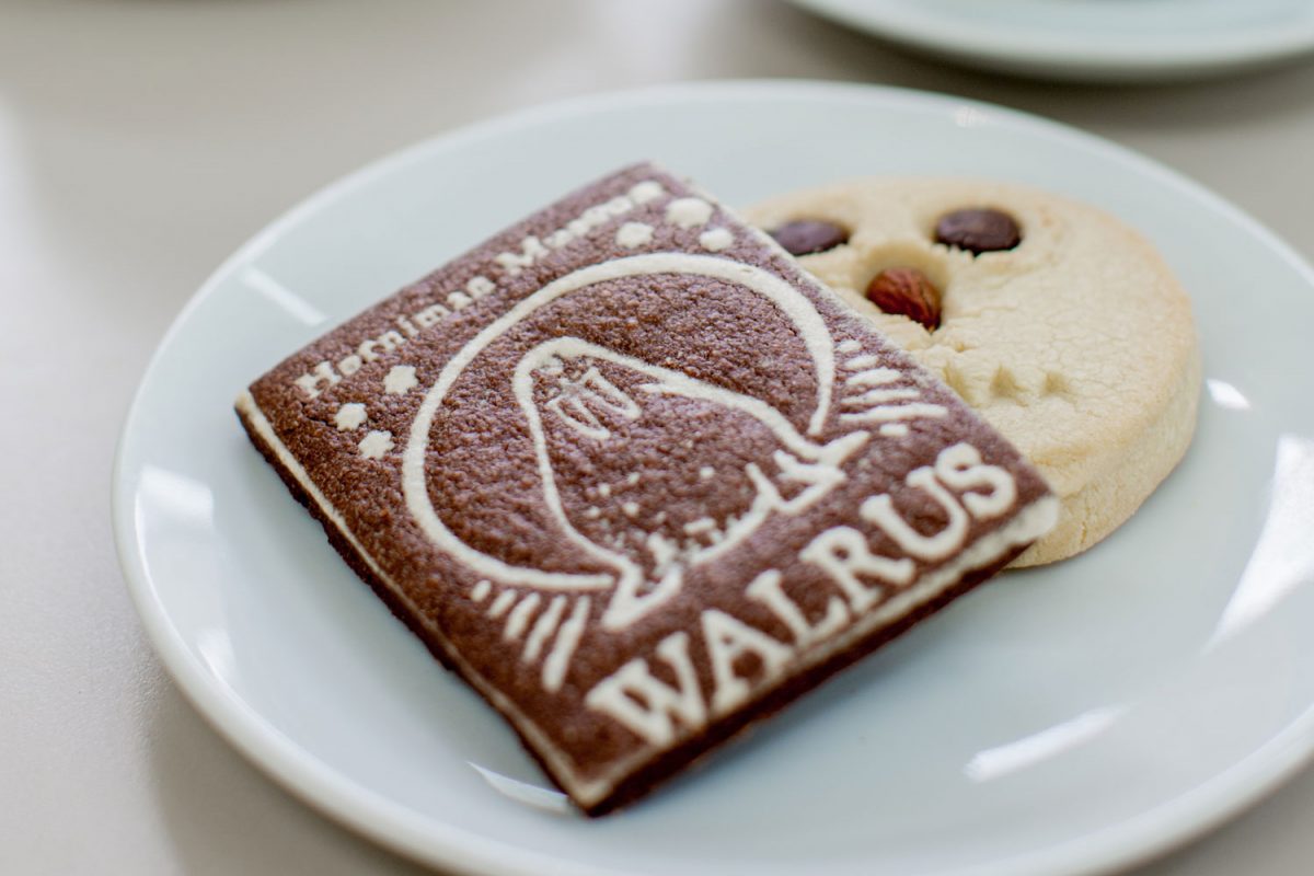 A plate with two biscuits next to two coffees in white china cups. One biscuit is shaped like an owl and the otehr had a picture of a walrus and the word walrus on it.