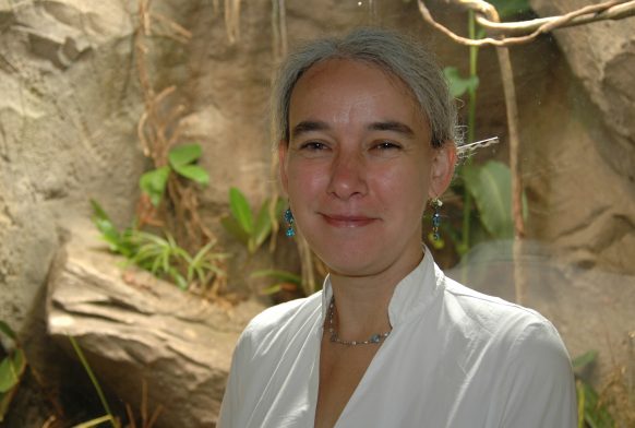 A woman is seen from the shoulders up stood in front of a rocky wall with a few climbing plants on it. She is smiling and wearing a white shirt
