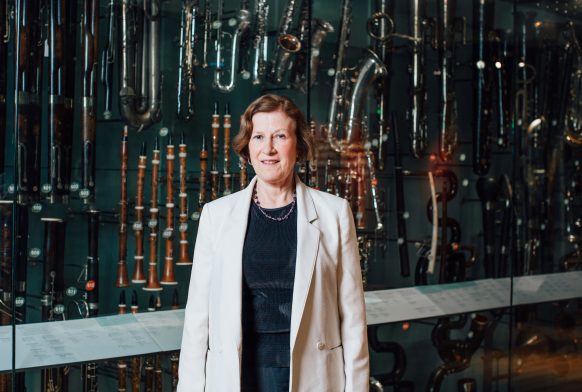 A woman in a white jacket is standing in front of a case with many recorders and other wind instruments on display in it
