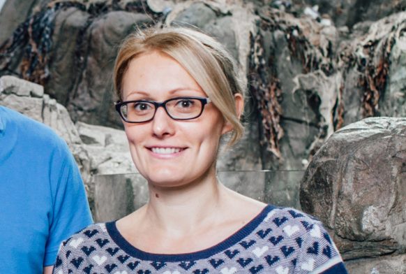 A woman in a blue striped top is seen from the thigh up. She is smiling and leaning on some rocks, which continue behind her with seaweed on them. Next to her is just seen a person wearing blue