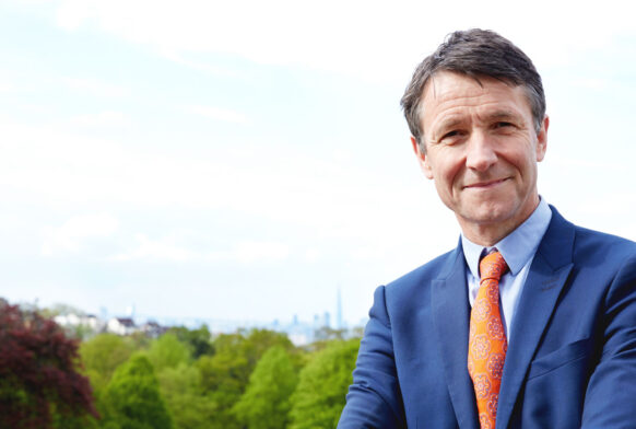 A man in a blue suit standing outside of a hill overlooking a city skyline with threes in the background. There is a structure to the right with windows all around and tiled roof.